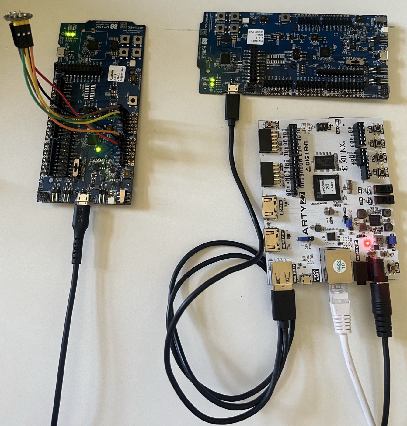 Three development boards on a white surface. Left: an nRF5340-DK with a microphone module wired to its header by coloured jumper leads, green LEDs lit, a micro-USB cable leaving the bottom. Top right: a second nRF5340-DK with green LEDs lit and a micro-USB cable in its nRF5340 USB port. Bottom right: a white Digilent Arty Z7-20 board with a Zynq-7000 chip, the black USB cable from the second nRF board plugged into its type-A USB HOST socket, a white Ethernet cable, and a barrel-jack power lead, with a red LED lit.